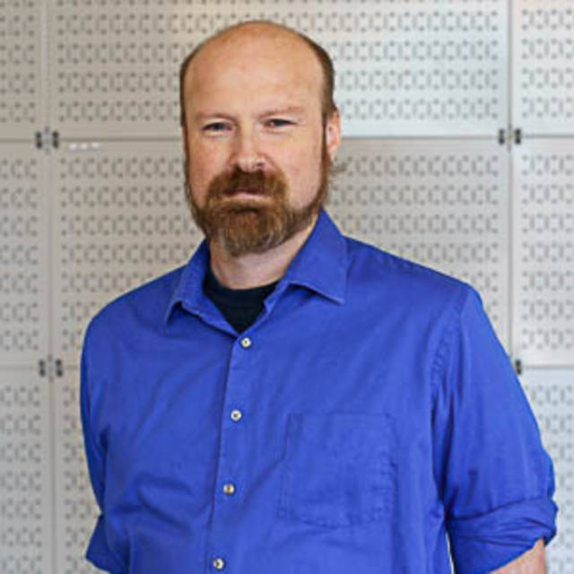 A person with a beard and mustache is wearing a blue shirt and standing in front of a patterned wall. The individual appears to be indoors.
