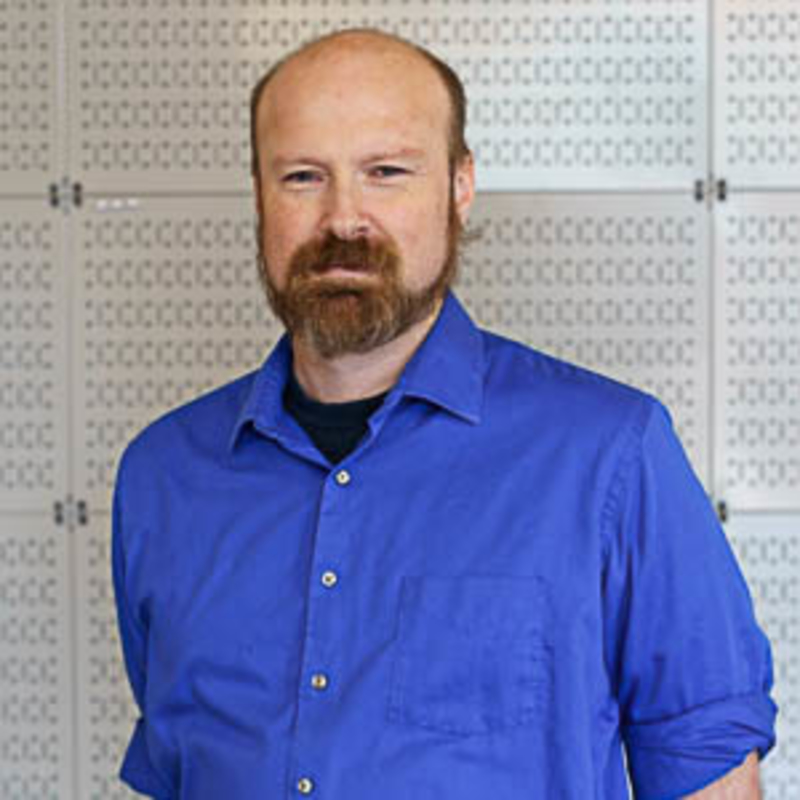 A person with a beard and mustache is wearing a blue shirt and standing in front of a patterned wall. The individual appears to be indoors.