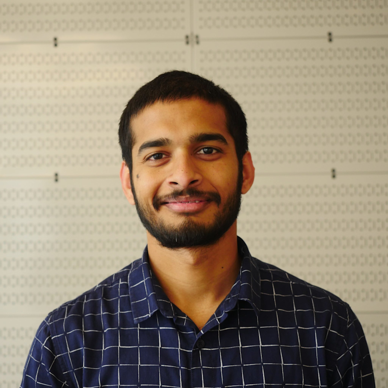 A person with short dark hair and a beard is smiling, wearing a blue checkered shirt. The background features a patterned wall.