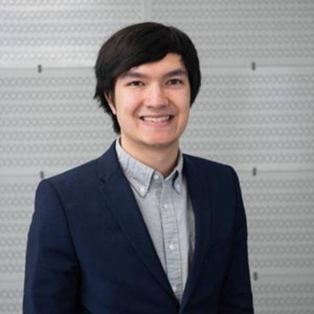 A person with short black hair and wearing a dark blazer over a light button-up shirt stands in front of a textured, light gray background, smiling at the camera.