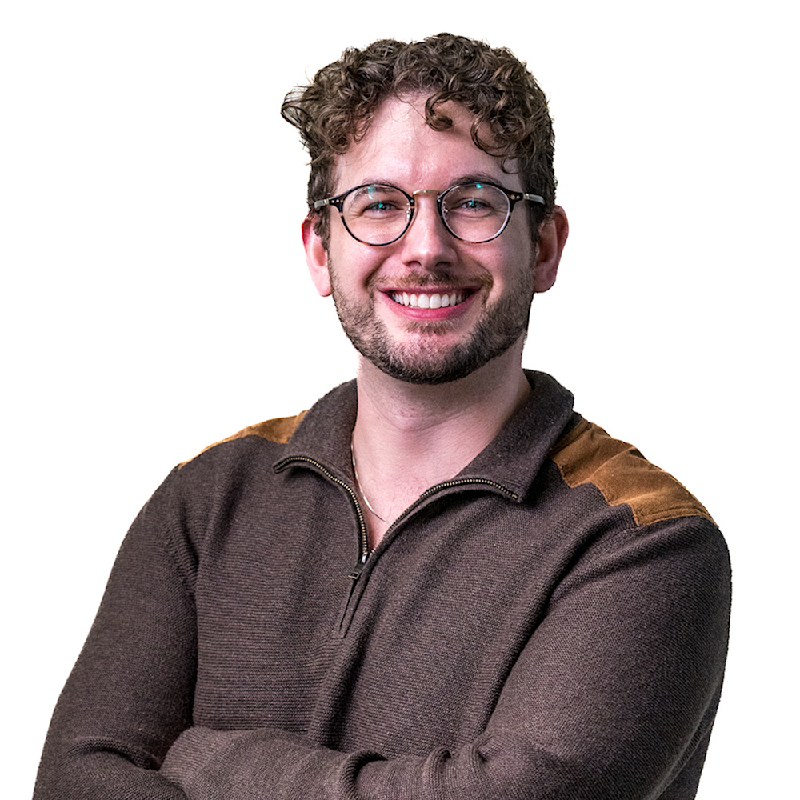 Smiling person with curly hair and glasses, wearing a gray sweater, stands in front of a patterned wall.