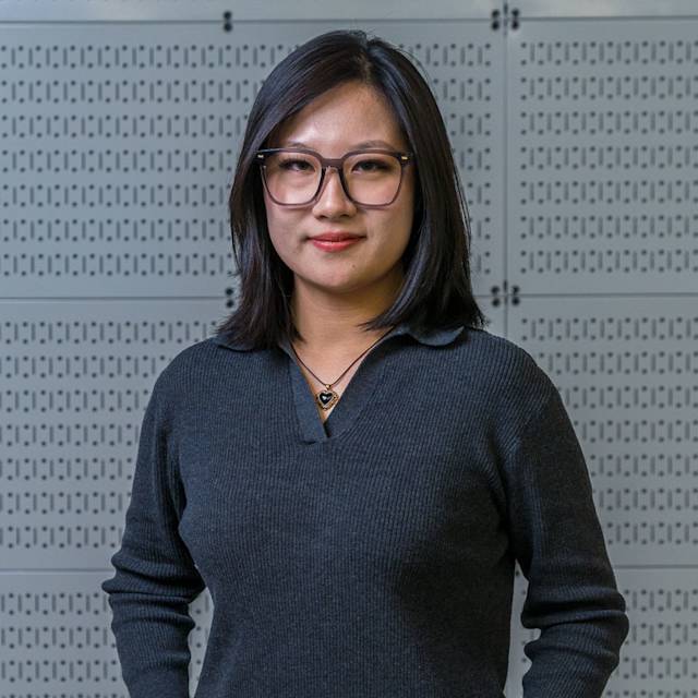 A person with shoulder-length dark hair and glasses stands confidently in front of a perforated metal panel. They are wearing a dark sweater and a necklace, smiling softly at the camera.