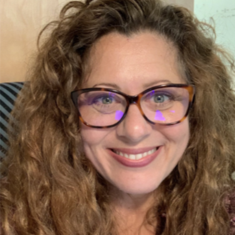 A person with long curly hair and glasses is smiling at the camera. Theyre wearing a dark top and are in an indoor setting.