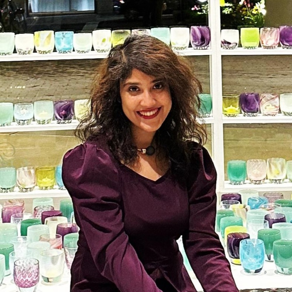 A person with long, wavy hair and a dark purple dress is smiling in front of shelves filled with colorful glass cups and candles. The cups are arranged in a variety of pastel shades.