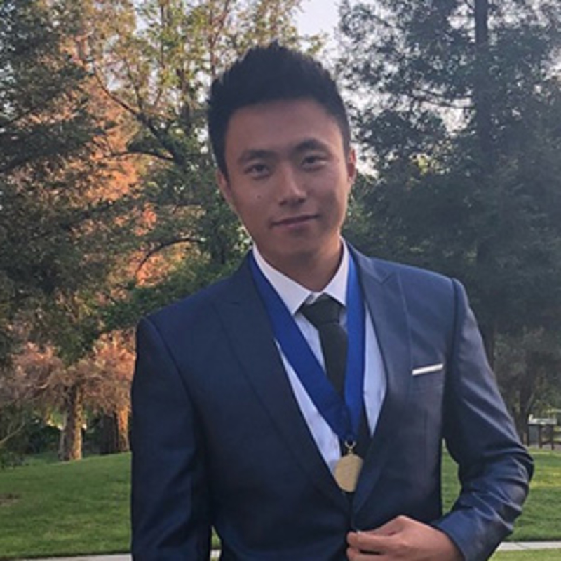 A person wearing a blue suit and tie stands outdoors, sporting a medal on a blue ribbon around their neck. Trees and greenery fill the background, and the individual is smiling slightly at the camera.