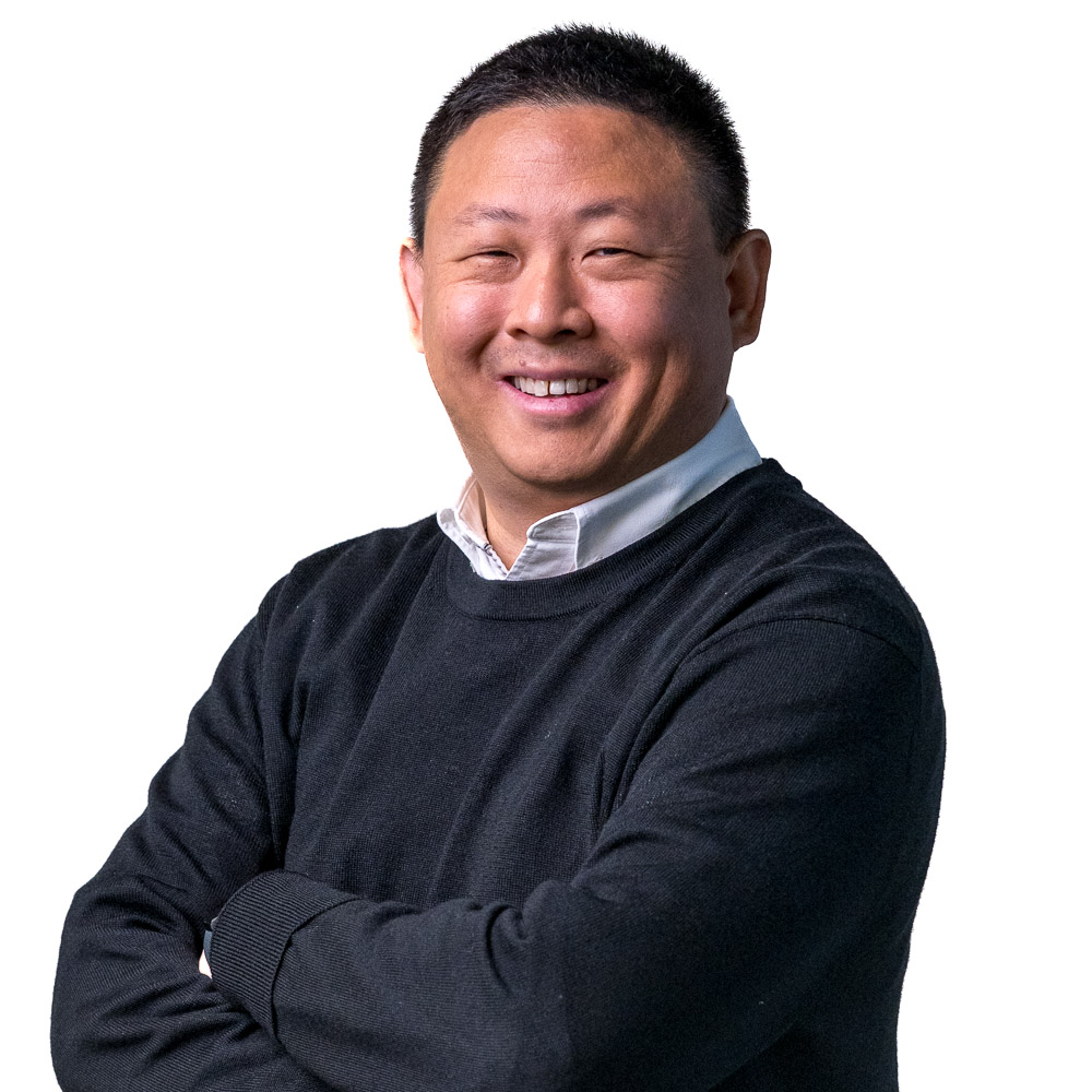 A person with short hair smiles while standing with arms crossed in front of a perforated wall. They are wearing a dark sweater over a light collared shirt.