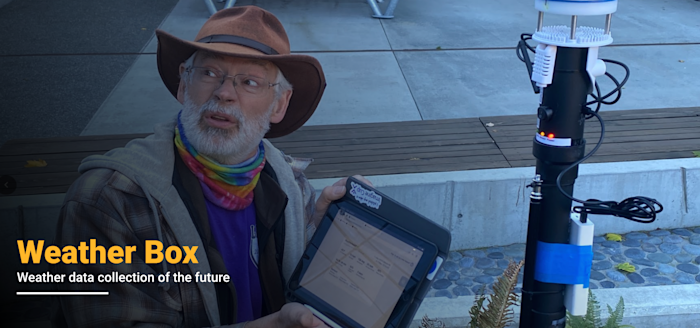 A person in a hat and colorful scarf holds a tablet displaying data. They are next to a tall weather instrument. Text on image reads: Weather Box - Weather data collection of the future. Background shows outdoor seating and paved area.