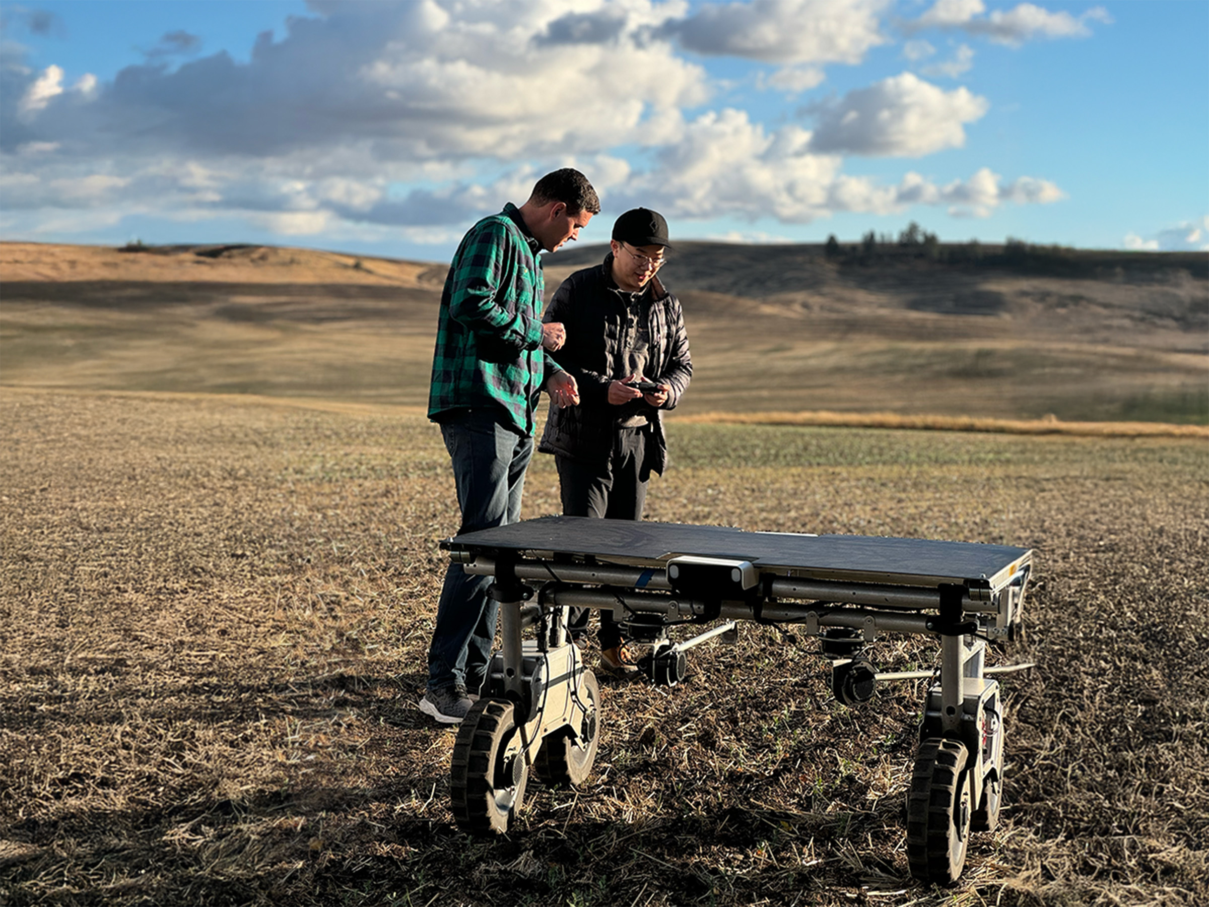 Two people examine a robotic vehicle in an open field under a partly cloudy sky. The terrain is arid and expansive, with rolling hills in the background.