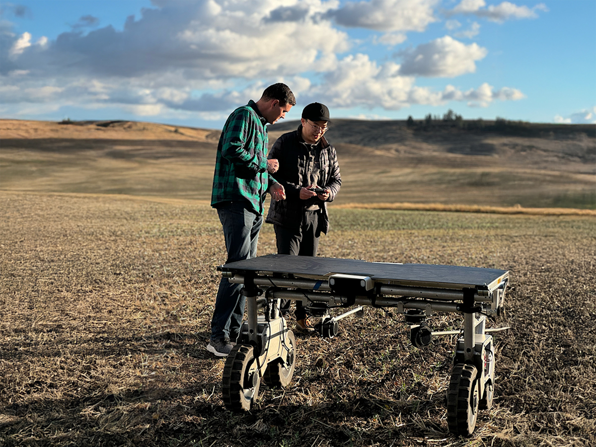 Two people examine a robotic vehicle in an open field under a partly cloudy sky. The terrain is arid and expansive, with rolling hills in the background.