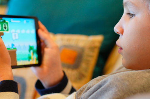 A child with blond hair is holding a tablet and playing a colorful video game, focused on the screen. Pillows are in the background, creating a cozy environment.