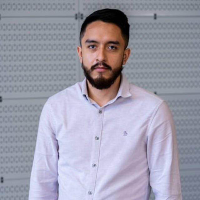 A man with a beard and mustache is wearing a light gray button-up shirt. He stands in front of a patterned, gray wall, looking directly at the camera with a neutral expression.
