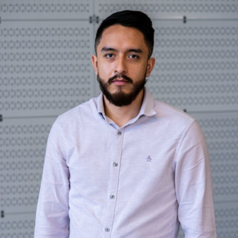 A man with a beard and mustache is wearing a light gray button-up shirt. He stands in front of a patterned, gray wall, looking directly at the camera with a neutral expression.