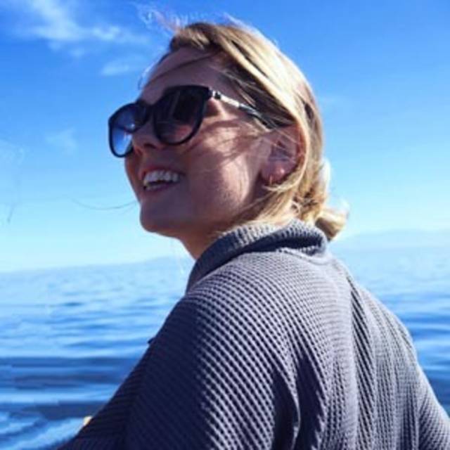 A woman wearing sunglasses smiles while looking to the right. She has blonde hair tied back and wears a textured dark jacket. The background features a clear blue sky and calm sea.