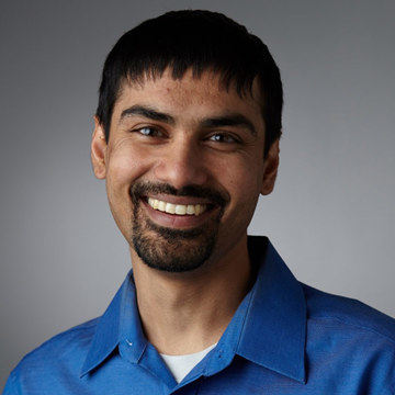 A person with short dark hair and facial hair is smiling at the camera. They are wearing a bright blue collared shirt and are set against a neutral gray background.