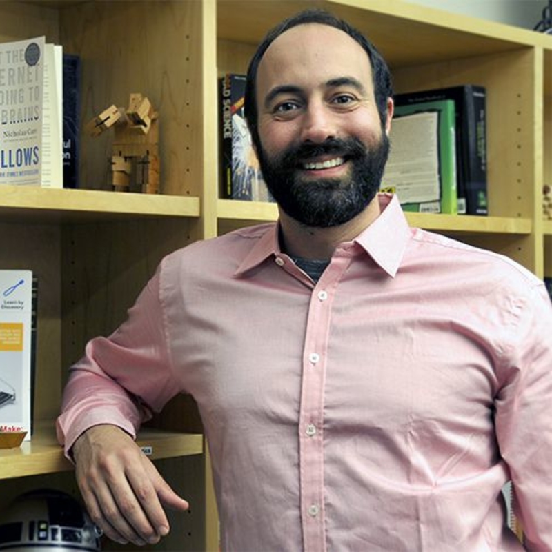 A person with a beard and smiling expression stands in front of bookshelves filled with various books and items. They are wearing a pink shirt and resting their arm on a shelf.