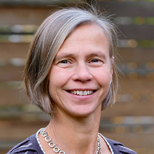 A woman with shoulder-length gray hair smiling in front of a blurred wooden fence background. She is wearing a necklace and a striped shirt.