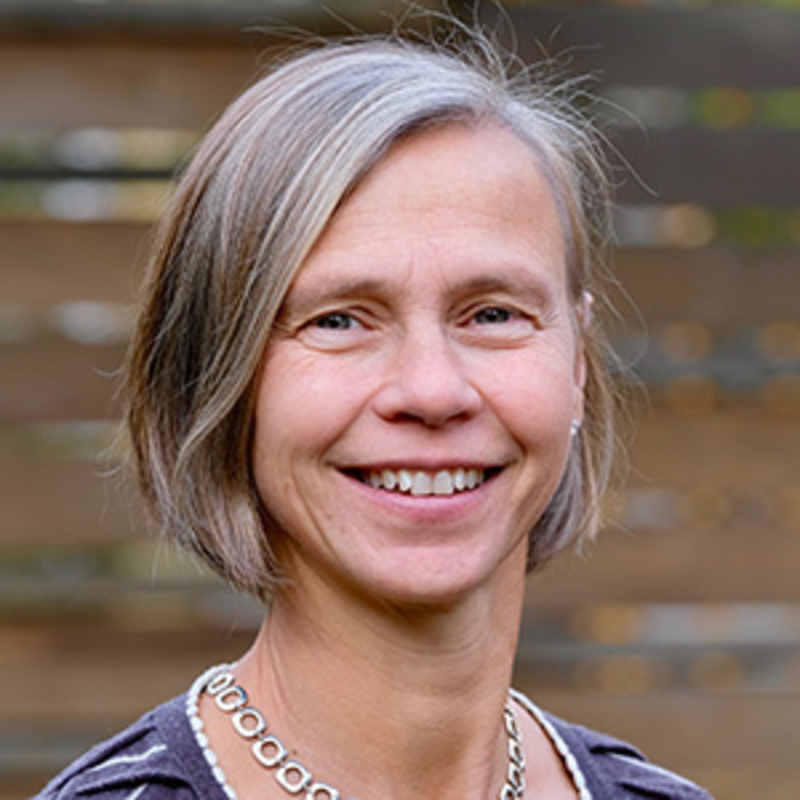 A woman with shoulder-length gray hair smiling in front of a blurred wooden fence background. She is wearing a necklace and a striped shirt.