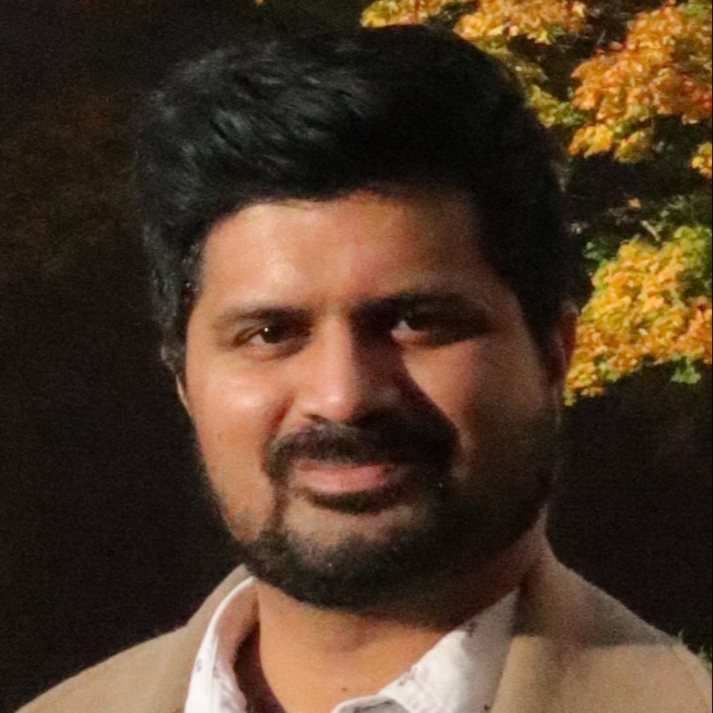 A person with short dark hair and a beard smiles at the camera. They are wearing a light-colored blazer and a white shirt. The background features autumn-colored leaves with shades of orange and yellow.