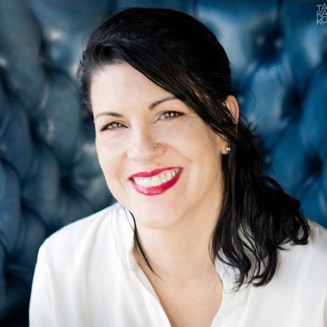 Smiling person with dark hair and red lipstick, wearing a white shirt, against a textured blue background.