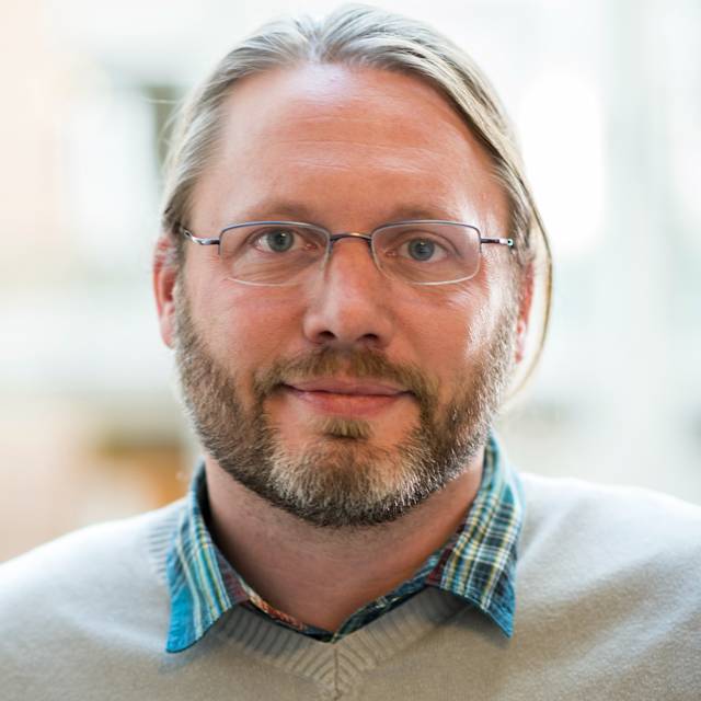 A person with glasses, light brown hair, and a beard is smiling slightly. They are wearing a plaid shirt under a light sweater. The background is softly blurred, suggesting an indoor setting.