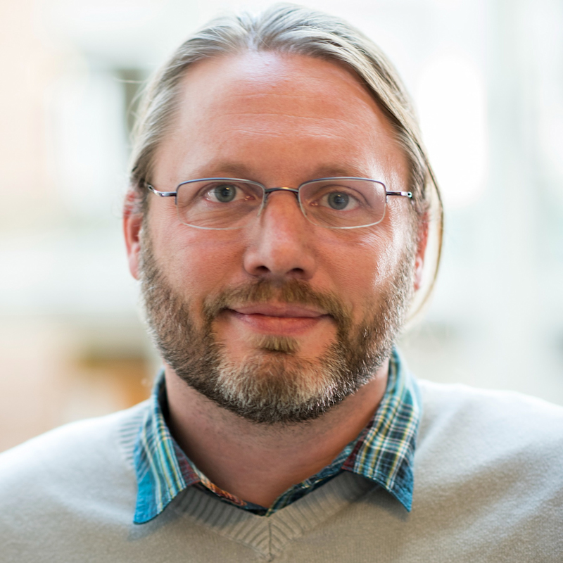 A person with glasses, light brown hair, and a beard is smiling slightly. They are wearing a plaid shirt under a light sweater. The background is softly blurred, suggesting an indoor setting.
