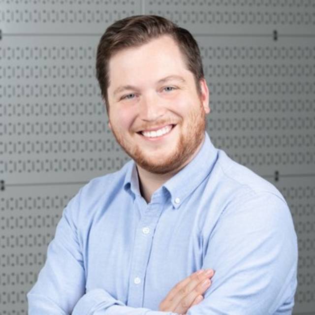 A man with short brown hair and a beard is smiling, wearing a light blue shirt. He stands with arms crossed in front of a patterned gray background.
