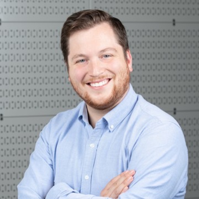 A man with short brown hair and a beard is smiling, wearing a light blue shirt. He stands with arms crossed in front of a patterned gray background.
