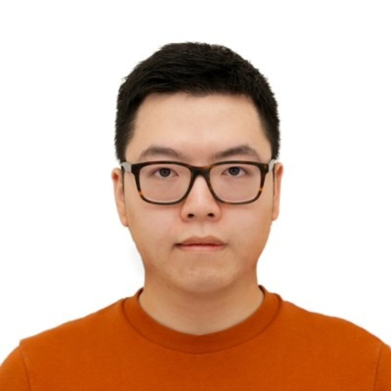 A person with short black hair, wearing black glasses and an orange top, is pictured against a plain white background, facing the camera with a neutral expression.