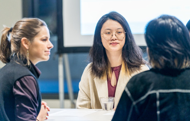 Three people are seated around a table engaged in conversation. One person is facing the other two, wearing glasses and a light-colored coat. There is a small glass of water on the table. A blurred screen is in the background.