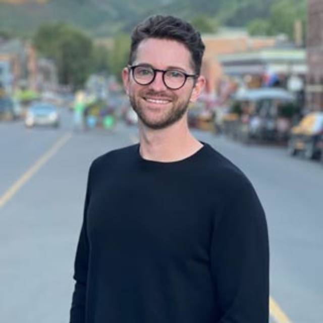 Man with glasses and a beard stands smiling on a street, wearing a black sweater. The background features a blurred view of people, shops, and greenery on a sunny day.