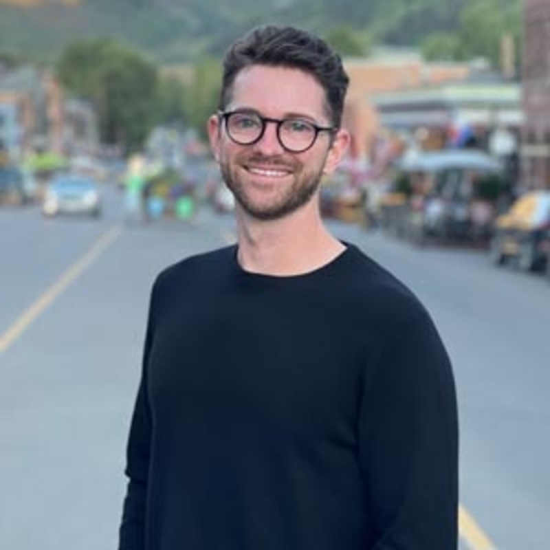Man with glasses and a beard stands smiling on a street, wearing a black sweater. The background features a blurred view of people, shops, and greenery on a sunny day.
