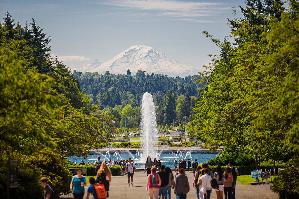 A scenic view shows people walking towards a tall fountain surrounded by lush greenery. In the background, a snow-capped mountain rises under a clear blue sky. The path is flanked by trees, creating a verdant and inviting atmosphere.