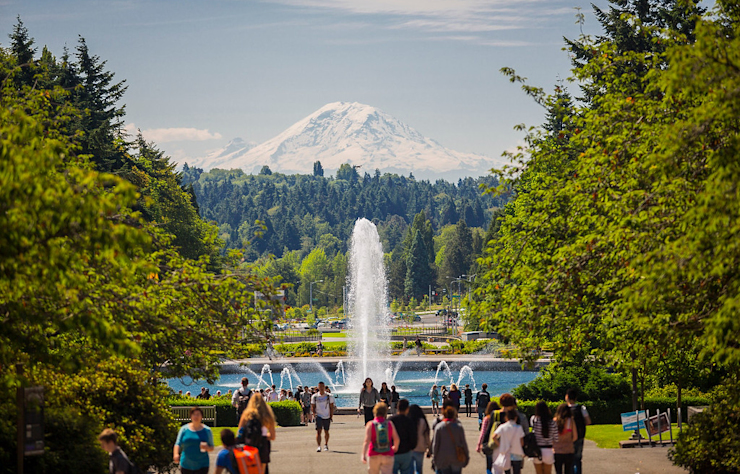 A scenic view shows people walking towards a tall fountain surrounded by lush greenery. In the background, a snow-capped mountain rises under a clear blue sky. The path is flanked by trees, creating a verdant and inviting atmosphere.