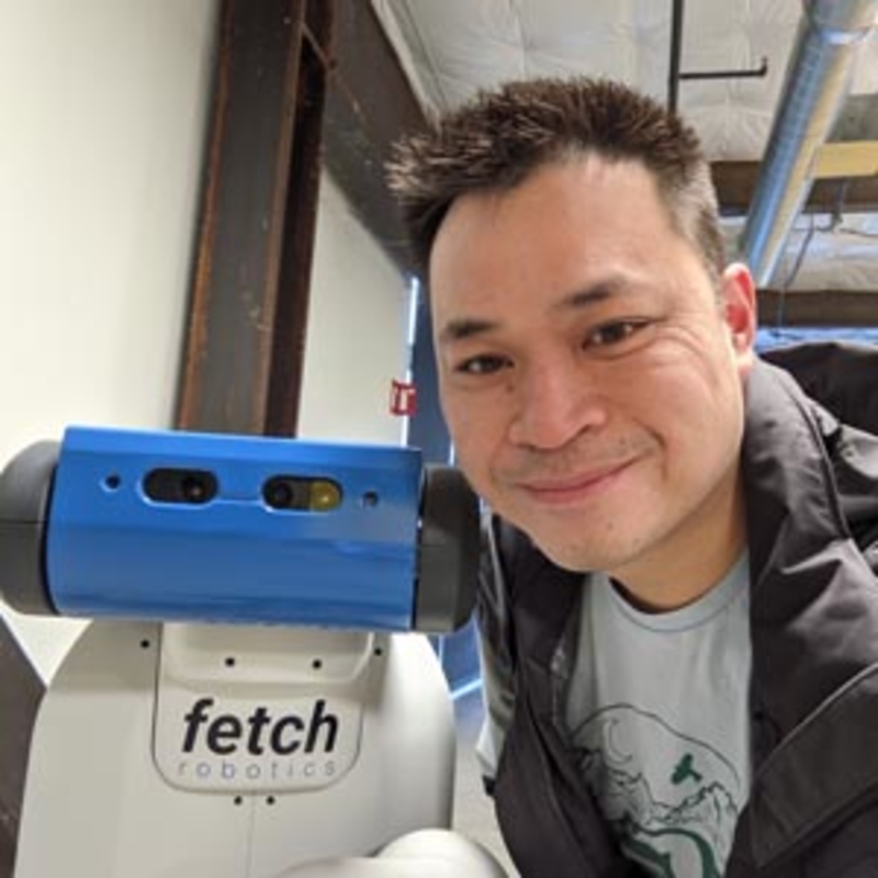A person smiling next to a blue and white robot labeled fetch robotics inside an industrial space with visible ceiling pipes. The person is wearing a dark jacket over a t-shirt, and the robot has sensors on top.