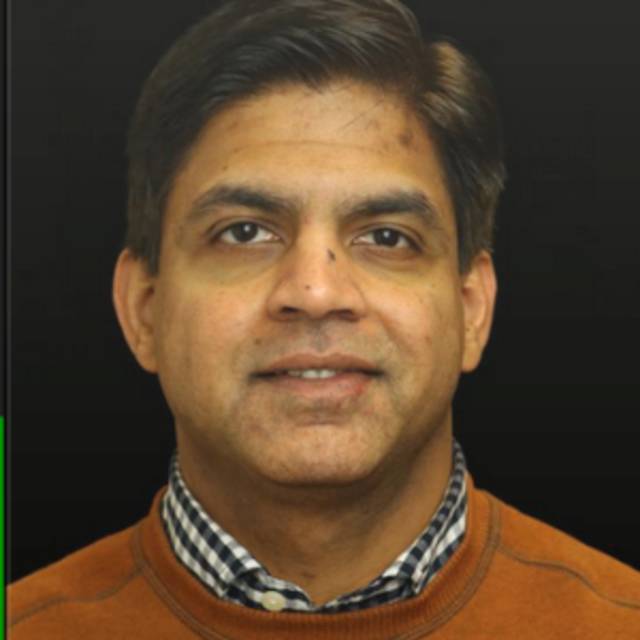 A person with short, dark hair is wearing a brown sweater over a checkered shirt and is smiling slightly against a dark background.