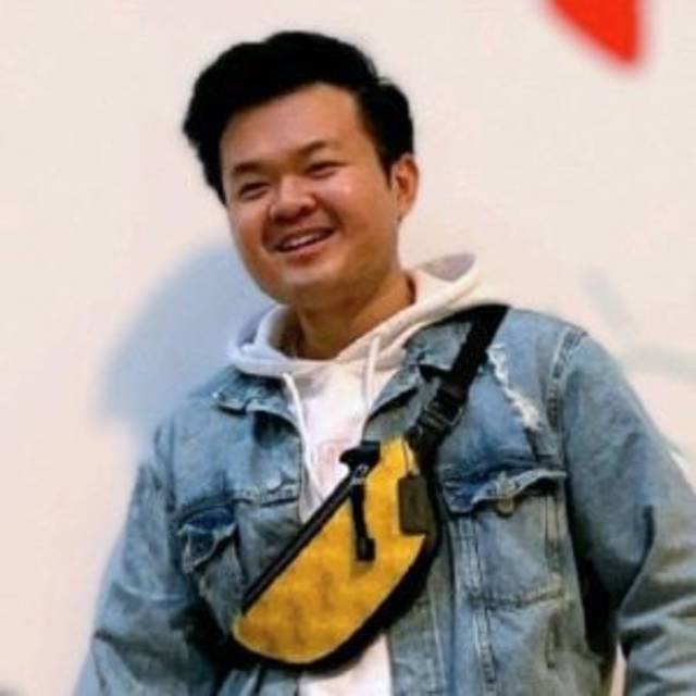 A person smiling while wearing a denim jacket over a white hoodie. They have a yellow crossbody bag with a black strap. The background is plain with a hint of red.
