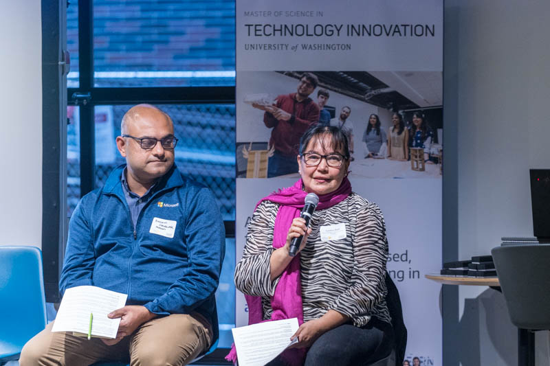 Two people speaking at an event. The man on the left wears glasses and a blue jacket, holding papers. The woman on the right, holding a microphone, wears glasses and a pink scarf. Theres a Technology Innovation banner from the University of Washington in the background.