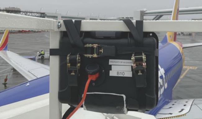 A piece of audio equipment is mounted on a platform overlooking an airport runway. The sky is overcast. An aircraft with a blue and yellow color scheme is visible in the background. An orange cable is connected to the equipment.