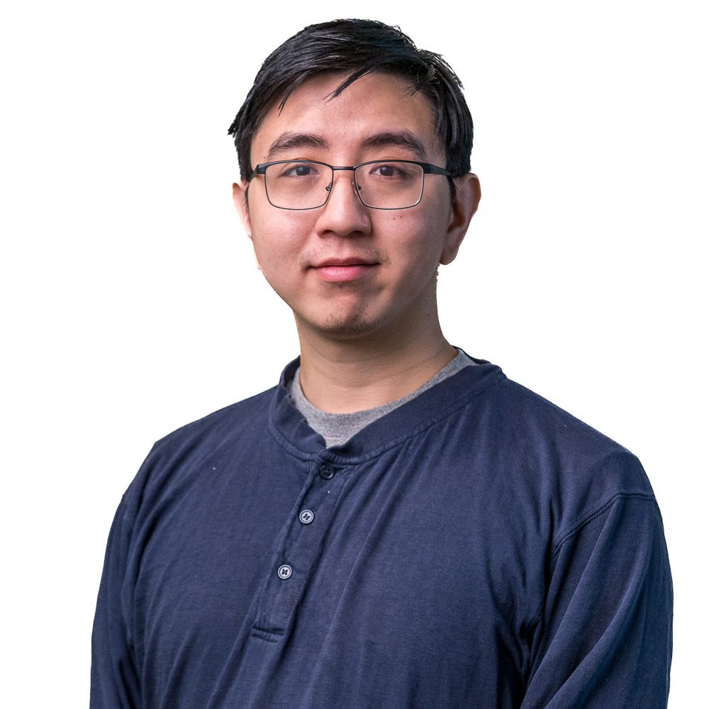 A person wearing glasses and a dark blue long-sleeve shirt stands with arms crossed, smiling at the camera. The background features a gray perforated wall.