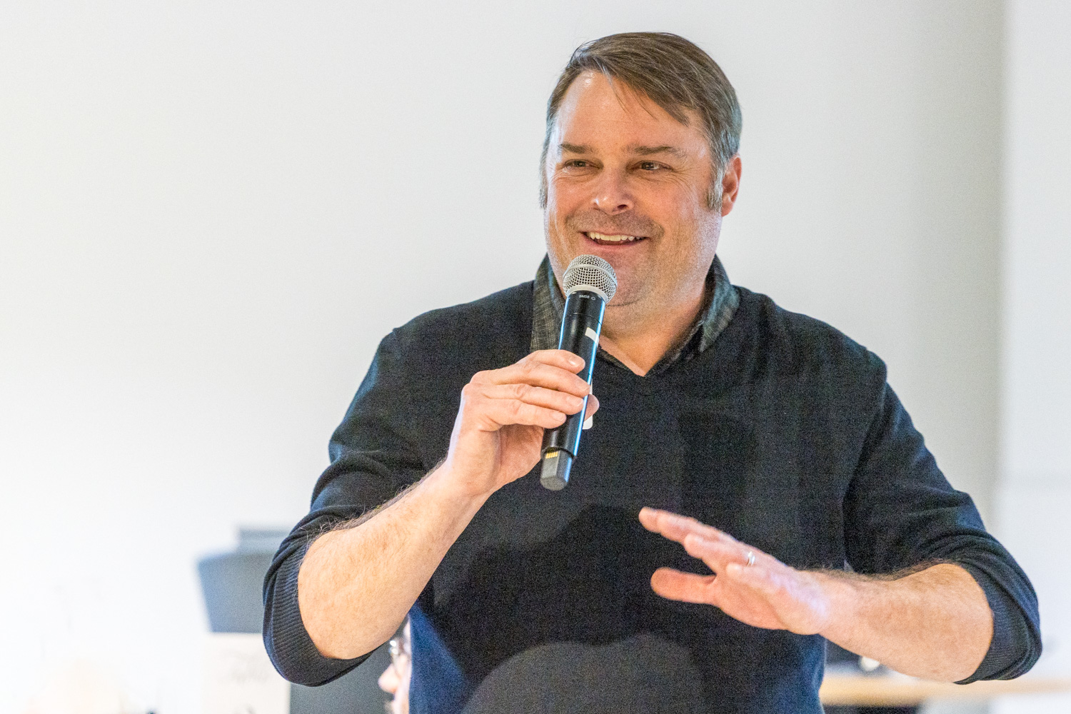 A man in a black sweater is smiling and speaking into a microphone, gesturing with his other hand. He stands against a plain, light-colored background indoors.