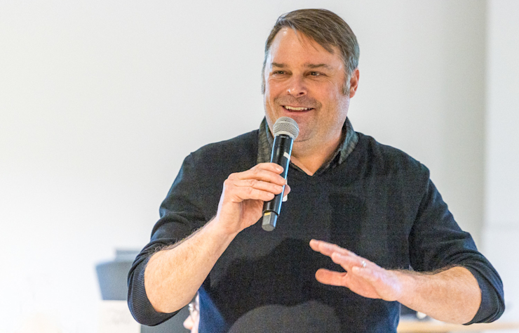 A man in a black sweater is smiling and speaking into a microphone, gesturing with his other hand. He stands against a plain, light-colored background indoors.