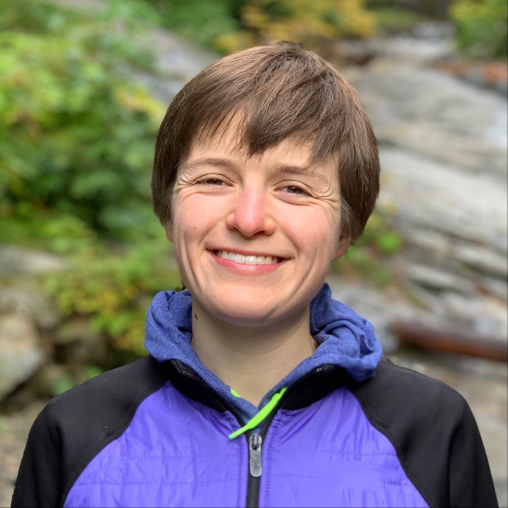A person with short hair wearing a blue and black jacket smiles outdoors. The background is a blurred natural setting with green foliage and rocks.