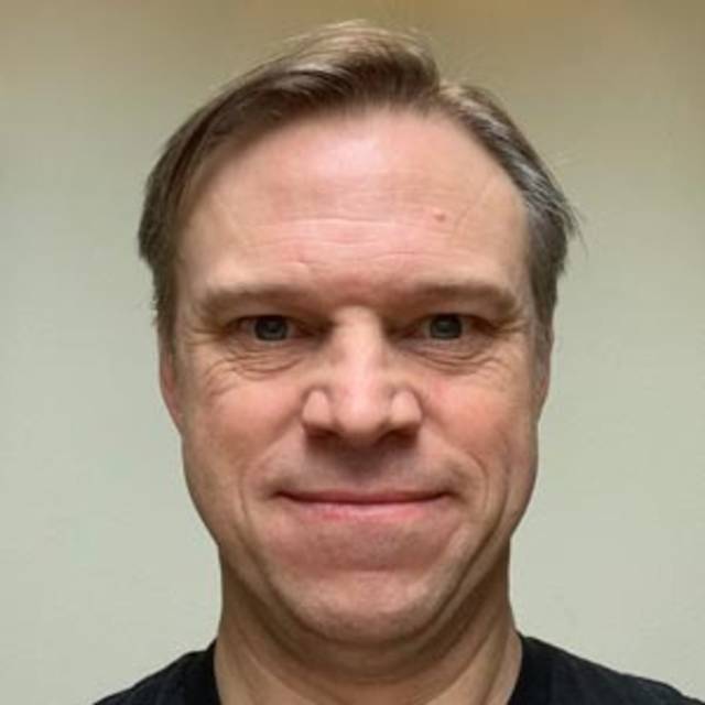 A person with short, light brown hair and a slight smile facing the camera. They are wearing a black shirt. The background is a plain, light-colored wall.