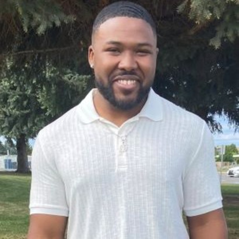 Man smiling, standing outdoors in front of a tree and grassy area, wearing a white short-sleeve polo shirt.