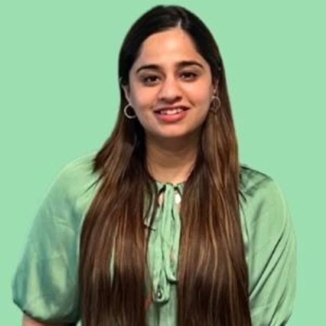 A woman with long, straight brown hair, wearing a light green top, stands in front of a solid light green background. She is smiling and facing the camera.