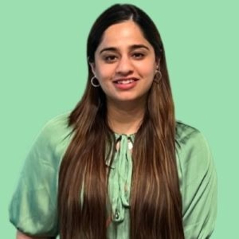 A woman with long, straight brown hair, wearing a light green top, stands in front of a solid light green background. She is smiling and facing the camera.