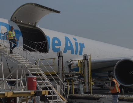 A cargo plane with Prime Air partially visible is being loaded. A person in a safety vest walks down stairs from the open cargo hold, and another person stands nearby. Loading equipment and a red container are in the foreground.