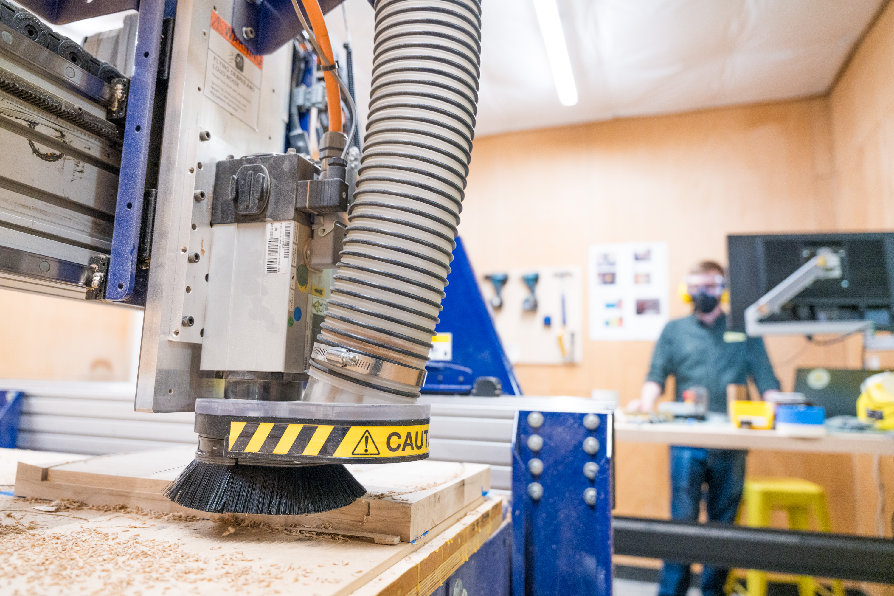 A CNC machine in operation, carving a piece of wood. The machine has yellow caution tape on the brush head. In the background, a person wearing a mask stands near a desk with tools and a computer monitor. The setting is a workshop.