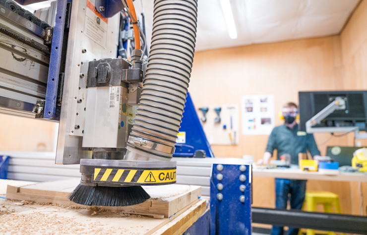 A CNC machine in operation, carving a piece of wood. The machine has yellow caution tape on the brush head. In the background, a person wearing a mask stands near a desk with tools and a computer monitor. The setting is a workshop.