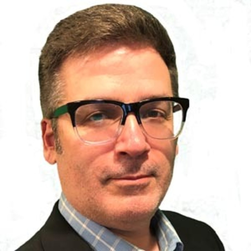 Man with short brown hair, wearing thick black glasses, a checkered shirt, and a dark blazer. He has a neutral expression and is facing forward against a light background.
